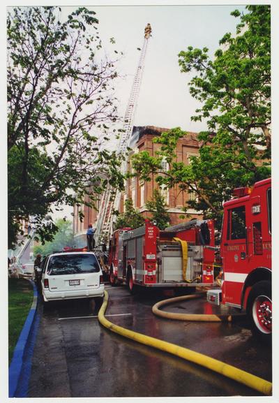 Fire trucks  and unidentified fire fighters are outside of the Administration / Main Building during the fire.  One fire fighter is spraying water onto the roof of the building from the top of a ladder that is towering over the building