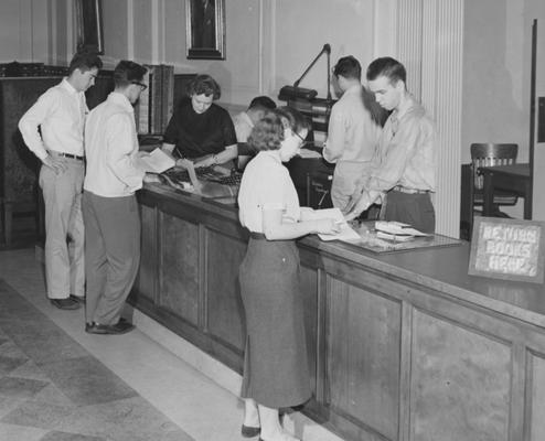 Circulation Desk--Circulation Desk--Great Hall, second floor; woman behind desk is Thelma Rogers, man behind desk (facing) is Ed Hall, man behind desk (back) is Ray Adams, and the woman on the other side of the desk is Sally Poundstone