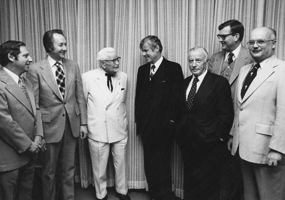 Seven men at the Sanders- Brown Center on Aging Building dedication. From left to right: Weck, unidentified man, Colonel Harland Sanders in white suit, Governor John Y. Brown, Jr., John Y. Brown, Sr., UK Vice President Ray Hornback, and Chancellor of Medical Center Peter Bosomworth. Received April 12, 1996 from the Medical Center Chancellor