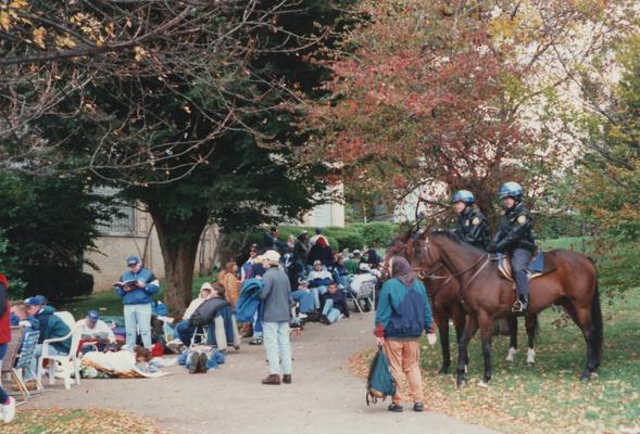 A color photo of unidentified people standing and sitting in line as well as two officers on horses at Memorial Coliseum for 