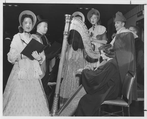 Six people singing while a woman plays a harp