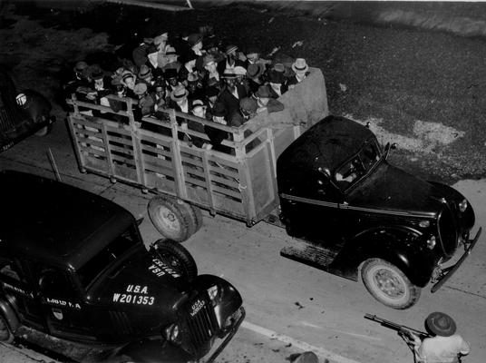 A crowd of miners in the bed of a truck, under guard