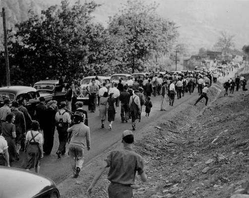 A crowd of men and women walking on a road with cars parked at the side