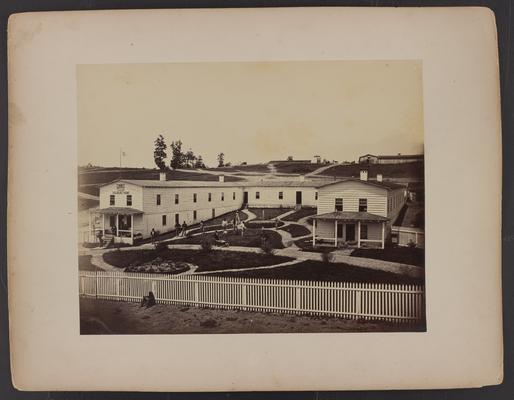 Exterior of building; large white wooden building, sign on building reads U.S. Sanitary Commission, Soldier's Home, large group of men standing in front, white wooden fence with men resting in foreground, dirt road leading to buildings in background