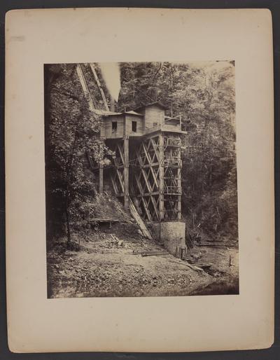 Steam powered pump house; wooden house built on mountainside raised with a concrete structure and wooden beams above the Kentucky River, three people looking out of window, men sitting on porch, trees and wooden staircase going over mountainside in background, creek in foreground