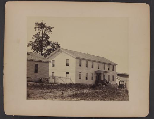 Exterior of building; large white wooden sided building flanked by smaller buildings, two African-American men in front, one on horseback