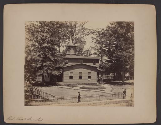 The Post Headquarters; wooden building with two floors and cupola, two men are standing on sidewalk near wooden fence in foreground, trees and house with wooden fence in background