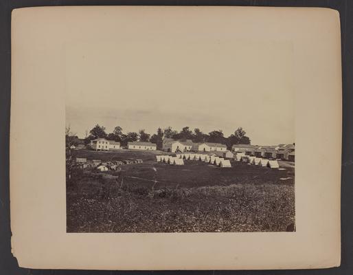 Landscape with building; three rows of white tents with wooden buildings behind them and two floor white wooden buildings adjacent, field in foreground and trees in background