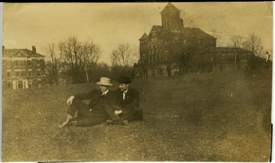 2 men sitting on the lawn of the University of Kentucky's campus with Barker Hall and Buell Armory  in the background circa 1914