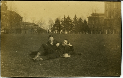 2 men sitting on the lawn of the  University of Kentucky's campus with Barker Hall, Buell Armory and the Administration Building in the background circa 1914