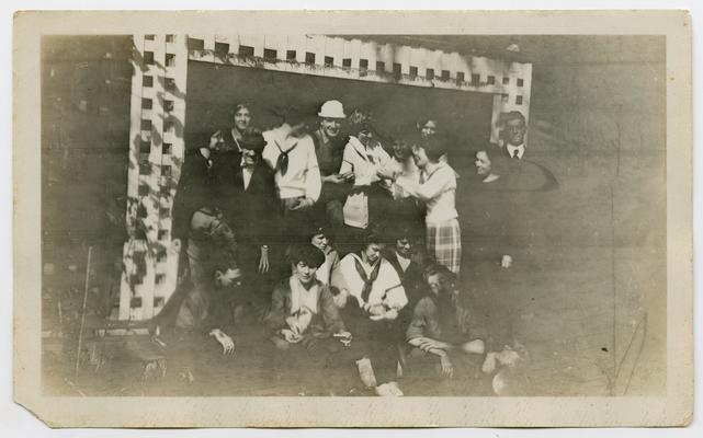 A large group of young men and women sitting and standing on the porch of a house or cabin.  Many of the women are wearing sailor shirts