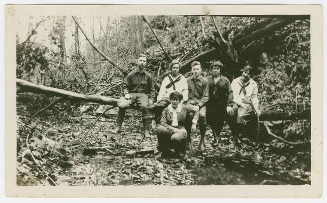 A group of four young women and two young men sitting on a log in a forest.  There is a pool of water in the foreground