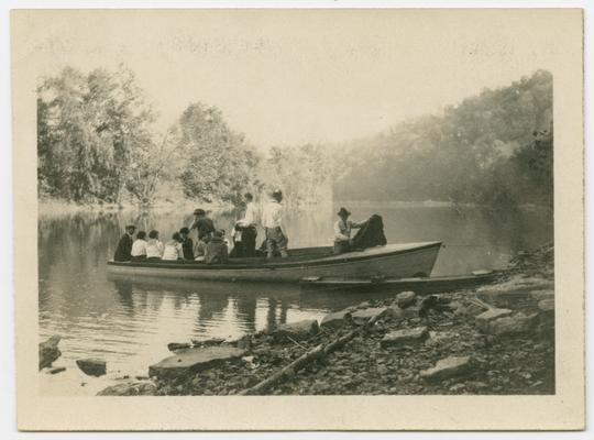 A group of men and women in a boat on the Kentucky River.  The foreground is a rocky beach.  The background is trees