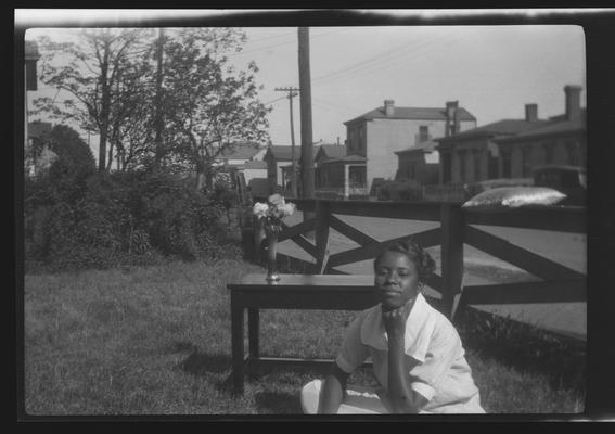 Negative of unidentified woman in yard next to a table with a vase of flowers on it