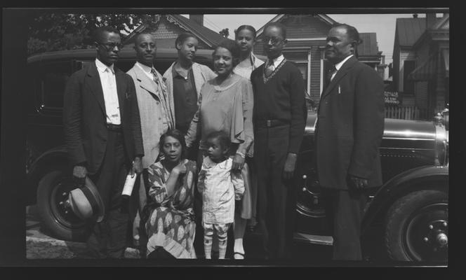 Negative of group of unidentified man, women, and a child in front of a car