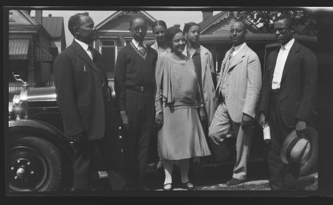 Negative of group of unidentified men and women in front of a car