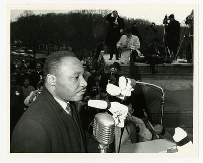 Dr. Martin Luther King, Jr., standing at a podium, photographers and crowd in background