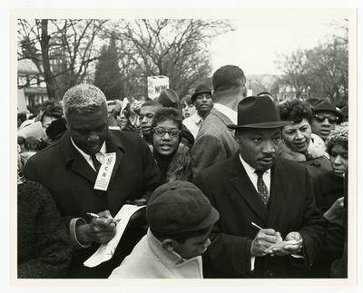 Dr. Martin Luther King, Jr., (right) and Jackie Robinson (left) taking notes during march, Georgia Davis Powers on extreme right over King's shoulder