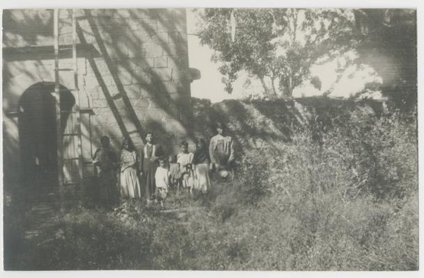 Eight adults and children stand in front of a stone structure, slightly to the left of an arched doorway.  The stone structure is probably the Catholic Church of Saint Catherine in Jalisco, Mexico. The majority of the people are probably Wixárika.  One of the adults is a Catholic priest.  There is a ladder leaning against the stone structure on the left