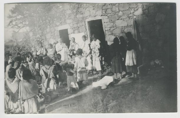 Twelve adults and children kneel on the ground and two people stand with their backs to the camera and facing six people with their faces toward the camera and standing in front of two rectangular doorways in a stone structure, probably at Ocota, Jalisco, Mexico.  The people are participating in a Catholic mass. The majority of the people are probably Wixárika.  Two of the people standing are Catholic priests