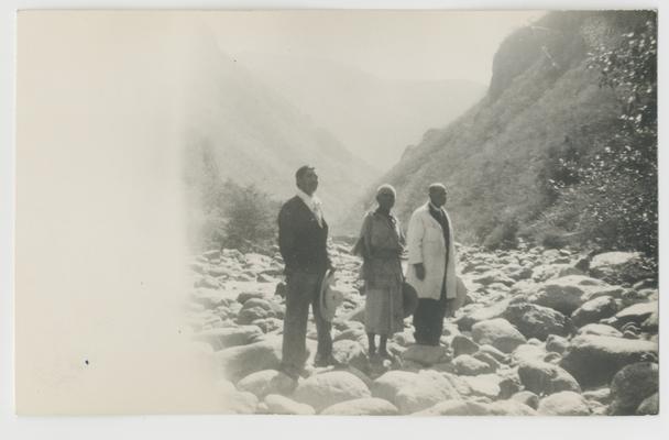 Three adults stand on a field of boulders with steep hills in the background at Barranca del Ríos de Chapalagana/Chapalagana River Canyon, Jalisco, Mexico.  One of these adults is the Governor of Santa Caterina/Saint Catherine, Jalisco, Mexico, and is probably Wixárika.  Two of the adults are Catholic priests