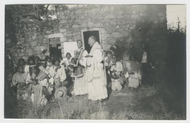 A large group of adults and children kneel and two adults stand in front of a ruined stone building with two rectangular doorways in San Sebastián/Saint Sebastián, Jalisco, Mexico. The building is probably a Catholic church, and the people are participating in a Catholic mass. The majority of the kneeling people are probably Wixárika. The two standing men are Catholic priests