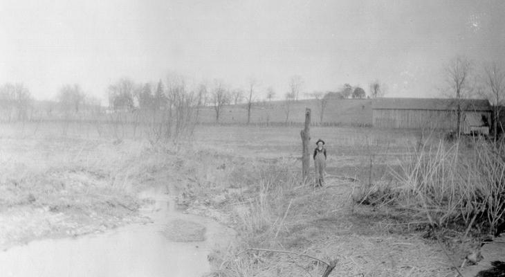 A view of the Right-hand Farm of Grassy Lick Creek taken from the bridge a short distance above the forks of Grassy Lick Creek. The direction is northeast, and the bottom beyond the boy standing by the post is where the 'Grassy Lick' was located in the farms of Grassy Lick Creek