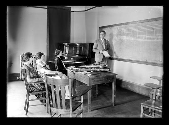 Class in music with Professor Lampert at blackboard, three women students, violin on the table