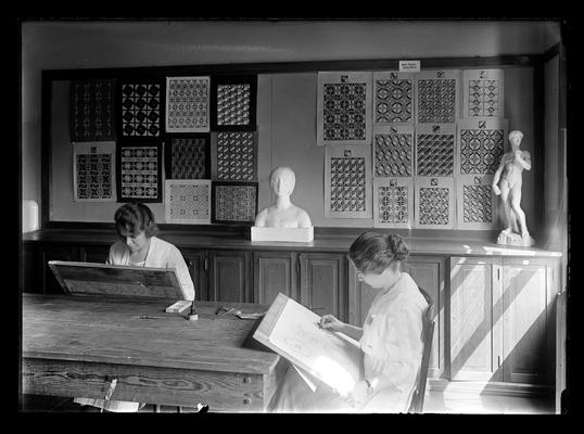 Two students sketching in an art class room, designs on wall in background, two pieces of sculpture on table