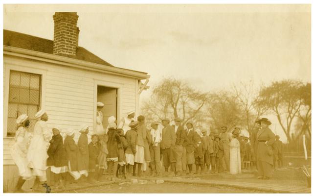 Maddoxtown students and Nannie Faulconer (1865?-1940) being served lunch outside the school house. Handwritten on verso, The penny lunch being served at Maddoxtown Colored School six miles from Lexington, Fayette County Kentucky. The majority of colored school in this county have regularly equipped kitchens. It is a windy day, Mrs. Nannie G. Faulconer, Superintendent inspecting