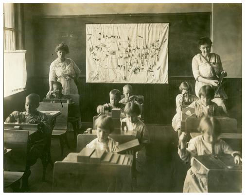 Picadome Montessori students working at their desks. Handwritten on oversized mat, Superintendent inspecting Montessori work in rural school Fayette County Kentucky. (Three copies, one Oversized)