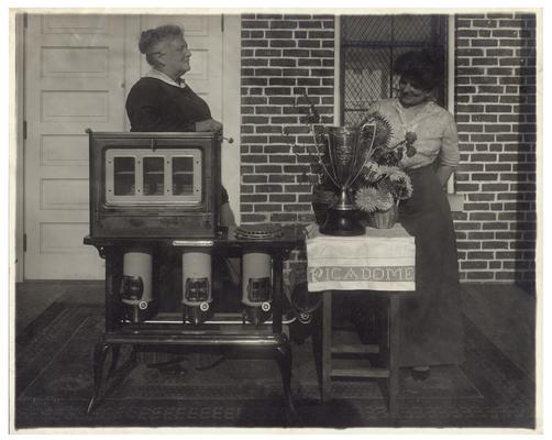 Portrait of Belle McCubbing and Nannie Faulconer (1865?-1940), posing with oven and trophy. Handwritten on verso, Silver Loving Cup and Blue Flame Oil stove won by Picadome rural school at the State Corn Show at Lexington, Fayette County Kentucky. The teacher Miss Belle McCubbing of Fayette County Ky. trained the class that won these prizes for the cheapest and best school lunch, and for the best soup made by the class after their own recipe