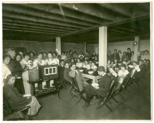 Group portrait of Picadome students eating lunch. Handwritten on verso, Hot nourishing soup is served everyday at this rural school. The children bring the greater part of the material for the soup pot. They are happy. The domestic science class makes the soup. Each child files by the soup pot, gets his portion and goes to his place. The girls take turn about in washing the dishes. The boys always take two bowls. (Two copies)