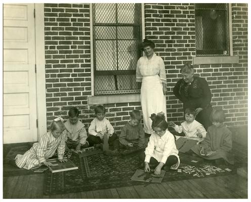 Picadome Montessori class doing work with Belle McCubbing while Nannie Faulconer (1865?-1940) looks on. Handwritten on verso, Class in Montessori taught by Miss Belle McCubbing at Picadome rural school near Lexington, Fayette County, Kentucky. The Superintendent, Mrs. Nannie G. Faulconer adopted for motto when she went into office many years ago the following expression, 'Progress, the onward stride of God.' by Hugo. (Two copies)