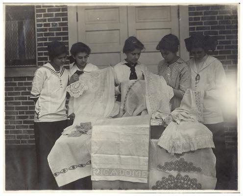 Group portrait of Picadome girls displaying sewing and needlework. Handwritten on verso, All Fayette County serving classes inspecting their finished work. This work has been done in the rural schools of Fayette county since 1909. The present Superintendent, Mrs. Nannie G. Faulconer has succeeded in the introducing Domestic Science, Needlecraft, Manual training and Home making into training schools of the Fayette County. (Three copies)