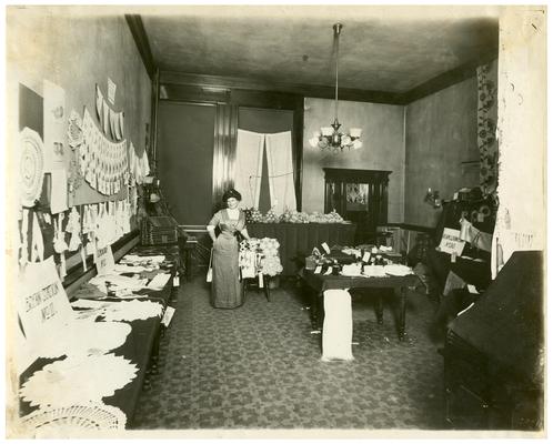 Portrait of Nannie Faulconer (1865?-1940) at the school fair with displays of students' work (A). Handwriting on verso. (Three copies, one in Oversize)