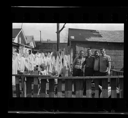 Four men behind fence, Woman on porch, Junk pile