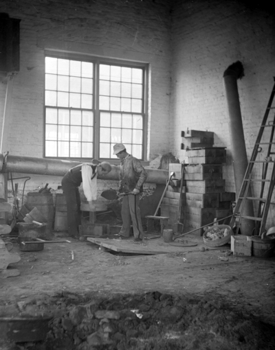 2 men working, one is holding a shovel and the other is bending over