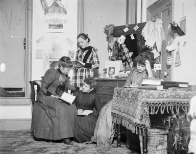 4 women together in a room, all are holding books, 2 of the women are sitting together talking