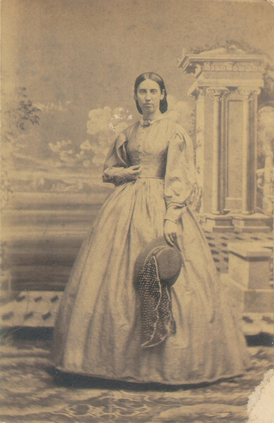 Woman in long dress holding hat, standing in front of painted backdrop