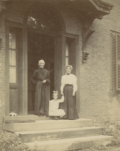 2 unidentified women standing, one is holding the hand of a young girl who is sitting on the stoop