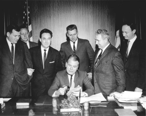 Kentucky Governor Bert T. Combs sitting at desk, future Governor Louie B. Nunn in black suit looking at camera, Len Press at far right (Frankfort, KY) (L to R: Bill Small, WHAS News Director; Ron Stewart, WBKY Chief Engineer; Harry King Lowman, Speaker of the House; Townes Ray, Majority Leader of the House; Wendell Butler, press)