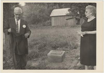 Man and woman standing near a marker on the ground (August 13, 1961). Pearl Day (Mrs. Wm. Everett) Bach is on the left. HGA Director, Mr. Henry A. Stovall, is on the back row, far right