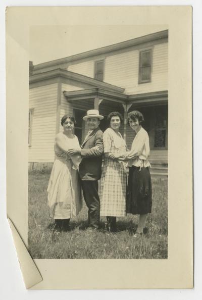 Two unidentified couples in yard of a house, one couple is a man and a woman, holding each other; the other couple is two women, holding each other