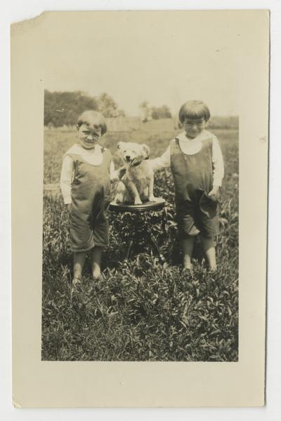 Two unidentified young children standing on either side of a dog sitting on a stool