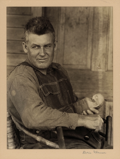 Bristol Taylor; Dulcimers, Farmer, Poet; Berea, Kentucky.  Man in overalls, seated in homemade chair, holding block of wood and carving tool