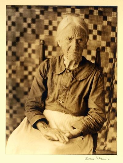 Miss Sarah Connelly; Weaver, Spinner; Brasstown, North Carolina.  Elderly woman seated in chair, with quilt hanging in background