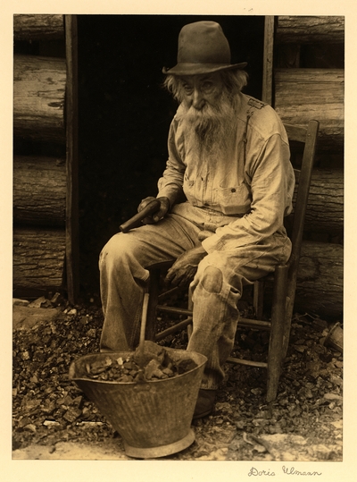 John B. Shell; Pine Mountains, Kentucky.  Elderly, bearded man in hat and patched pants, seated in chair in front of doorway; coal bucket? sitting in front of him