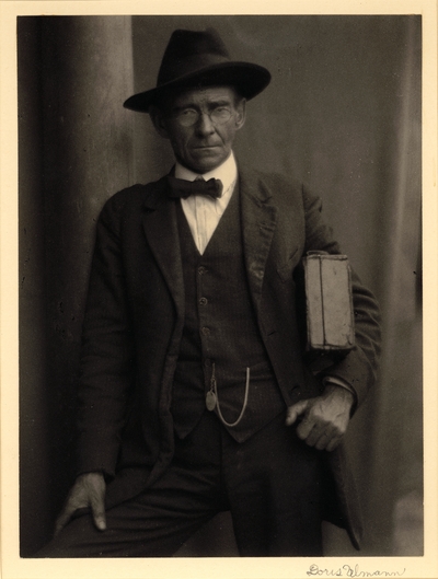 Preacher.  Man in hat, glasses, bowtie, and suit, standing with case under arm