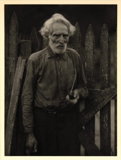 Nick Barton, Civil War veteran, d. May 1928.  Elderly, bearded man in polka-dot shirt, standing in front of gate, holding pipe
